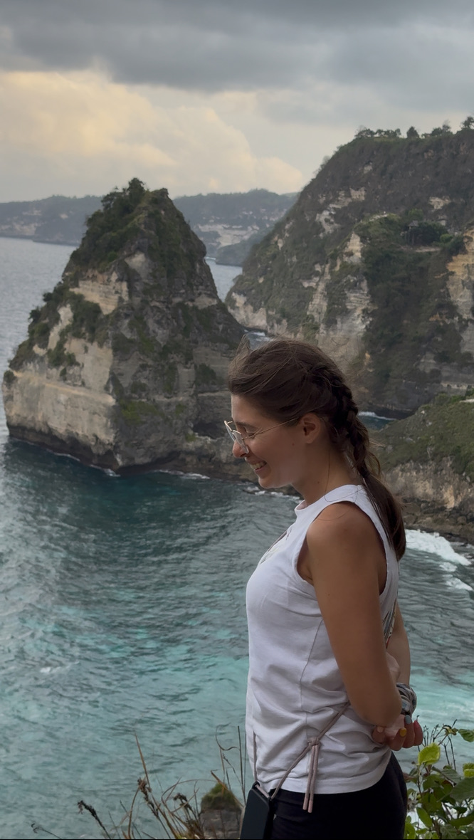 Anca standing relaxed at a dramatic clifftop viewpoint in Bali — white top, braid, looking calmly at the view, completely at ease