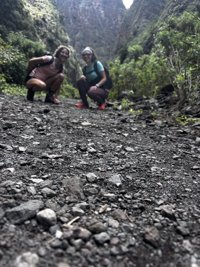 Anca and Giulia crouching on a dark volcanic gravel trail inside a narrow canyon gorge in Tenerife — the camera low to the ground, lush green canyon walls rising steeply behind them