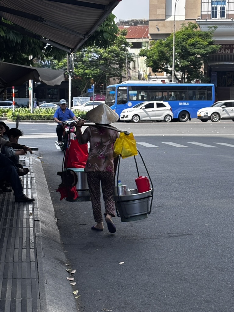 A Vietnamese woman in a traditional conical hat carrying food containers on a yoke through a busy Ho Chi Minh City street