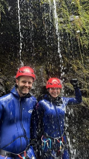 Anca mid-rappel on the waterfall face, water rushing directly over her, rope taut, total concentration